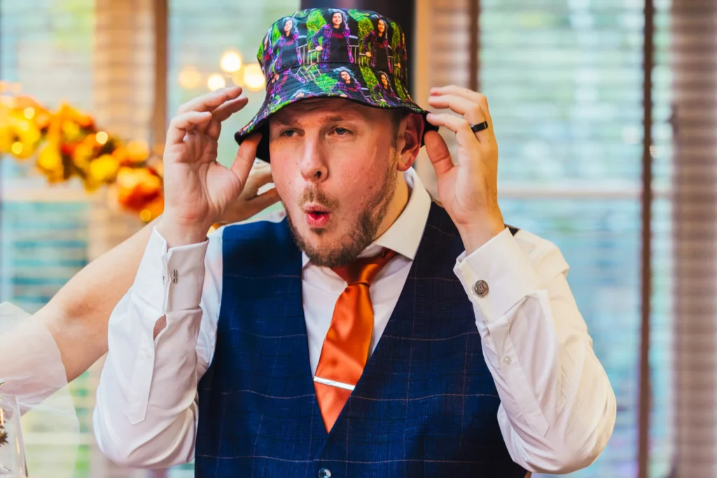 one half of the couple is wearing a hat with his partners pictures printed on it he is pulling a funny face as he pulls the hat down on his head