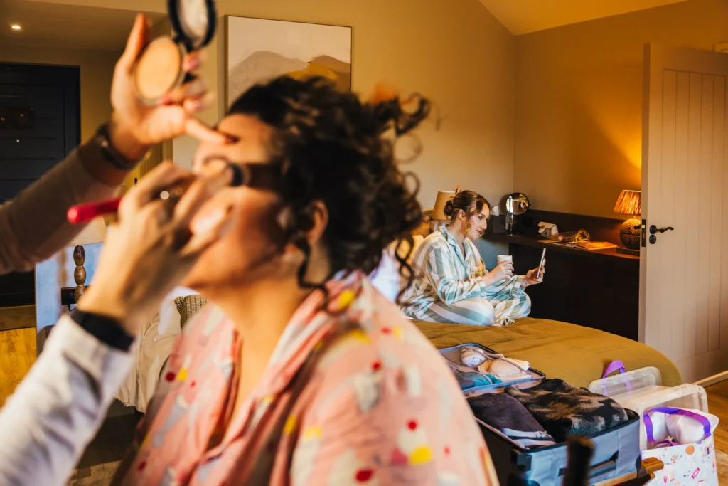 Two bridesmaids getting ready for a wedding one is in the foreground getting her make up done in her pjs and the other is in the background on FaceTime to her friend.