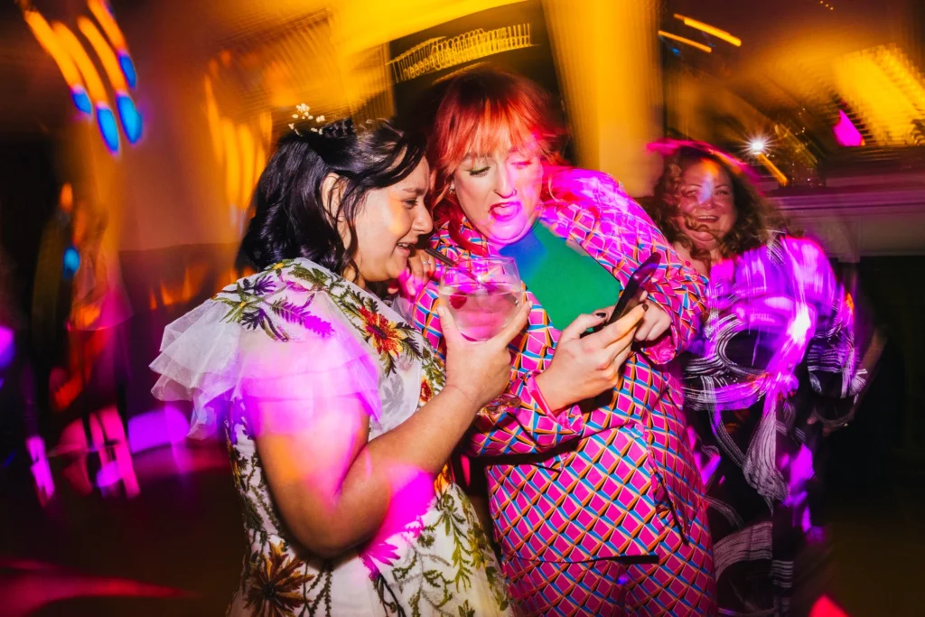 wedding guest is showing bride something funny on her phone, shes pulling a funny face whilst the bride laughs