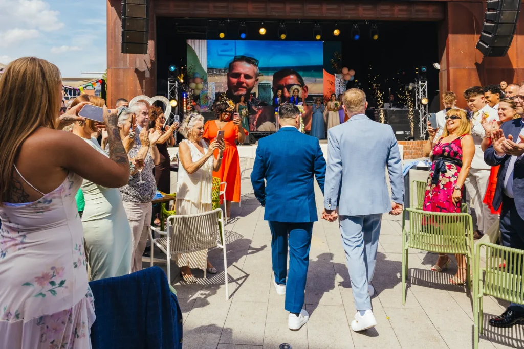 Couple walking to stage for their wedding ceremony, guests clap and film them walking down the isle and sparklers are going off. Their wedding party and drag queens are waiting on stage for them and there's a big picture of them on a screen