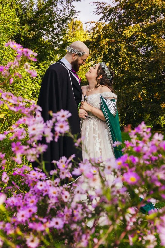 Couple portrait with lots of purple flowers in the foreground. Both are wearing capes and head pieces.