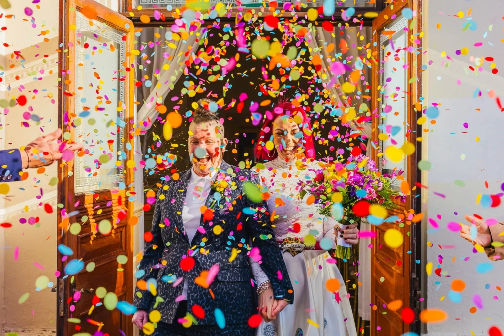 Couple leaving their wedding venue in a cloud of colourful confetti. They have the biggest smiles on their faces and look phenomenal one has a leopard print suit and the other wears a white dress with a pink belt.