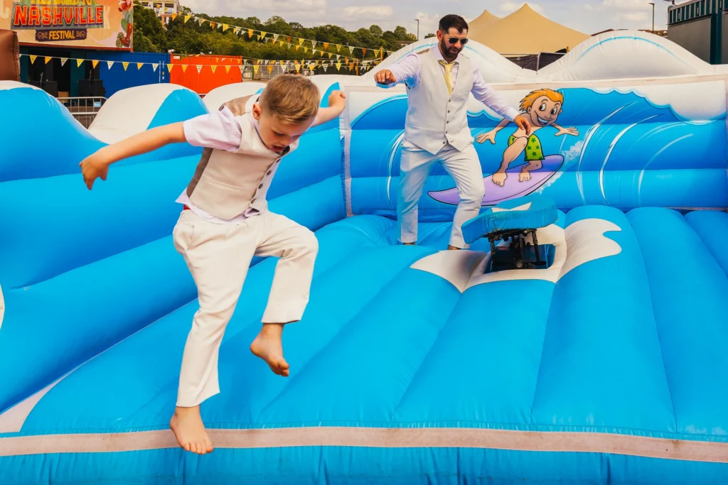Little boy jumps off of bouncy surf board game at wedding whilst someone else gets ready to take their turn behind him.