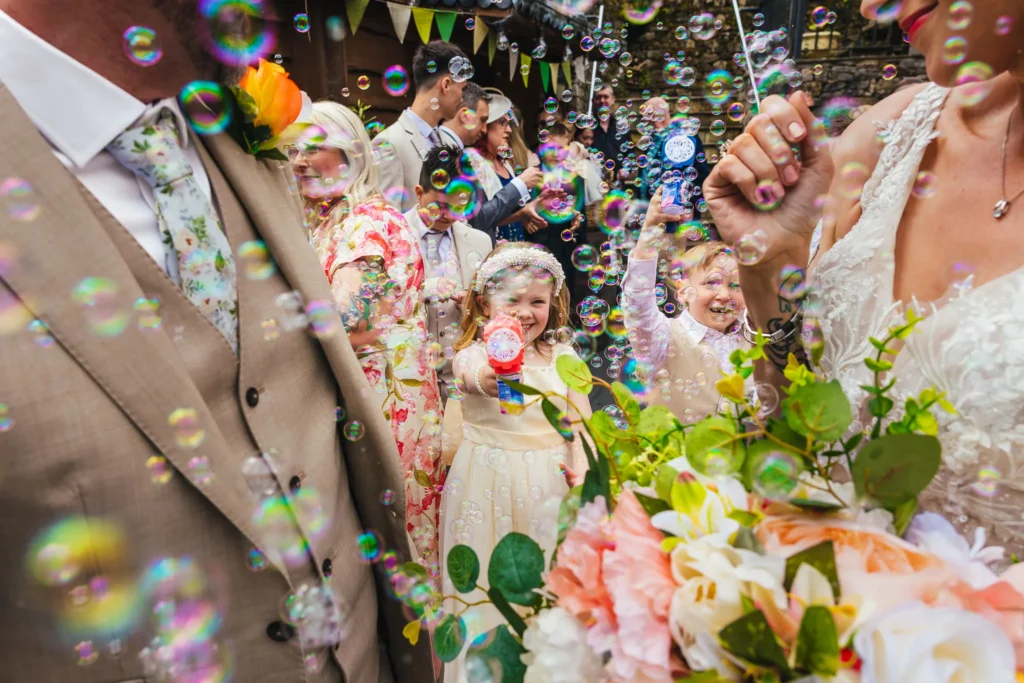 Wedding children blowing bubbles at couple using a bubble wands. Couple are both looking back at them with smiles