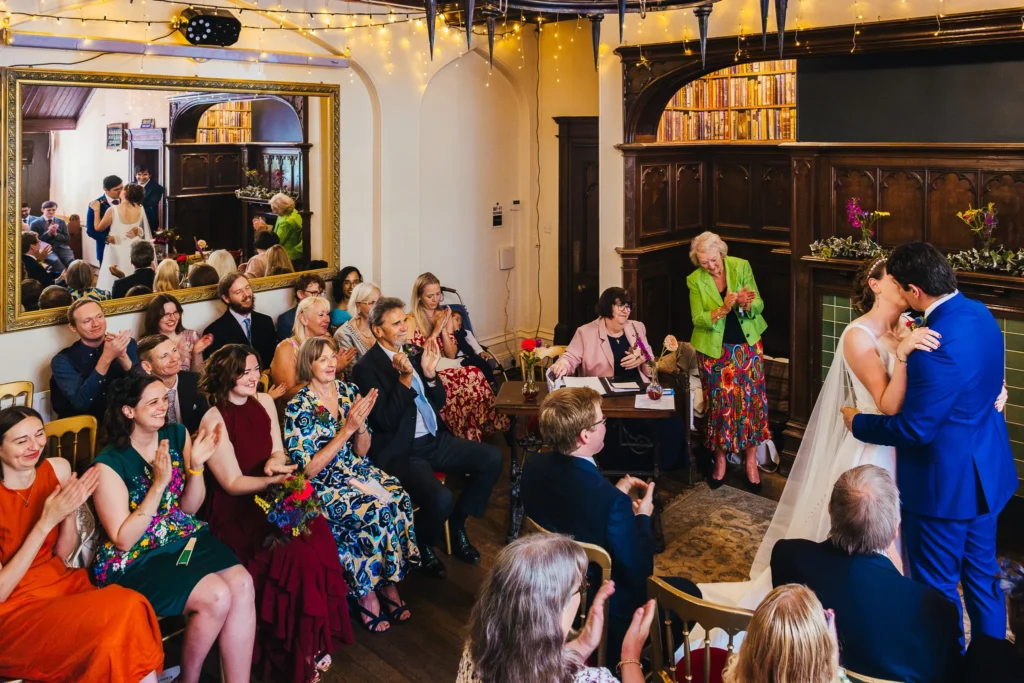 Couple sharing first kiss at the end of their wedding ceremony. You can se all their guests clapping as well as the couples reflection in the mirror on the wall.