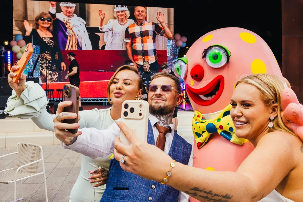 Mr blobby takes selfie with three guests during wedding cocktail hour. Whilst the groom and best person are in photo form behind with the king and queen