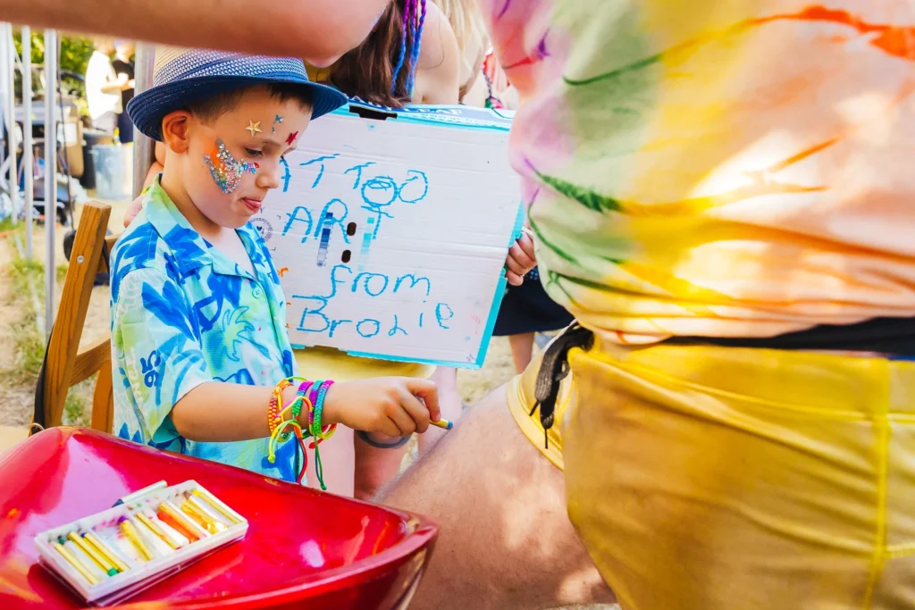 Little child does washable tattoo art on guest wearing gold hot pants and tie dye tee. Child is wearing a blue hat and shirt with glitter on his face.