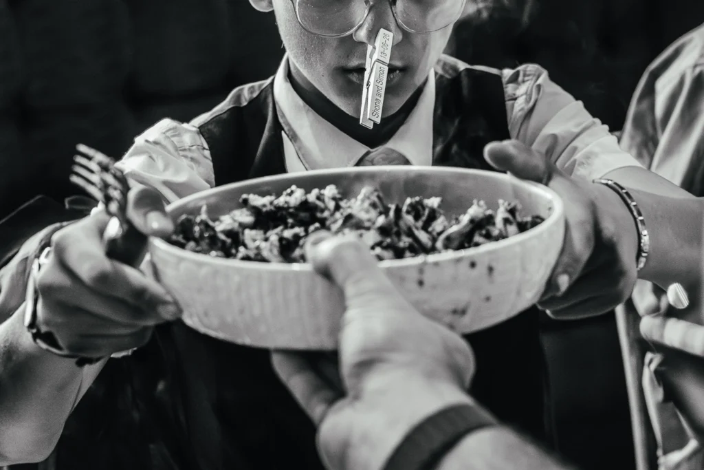 Guest being handed food during wedding breakfast they have a peg on their nose with the couples name and wedding date on it. The image is black and white.