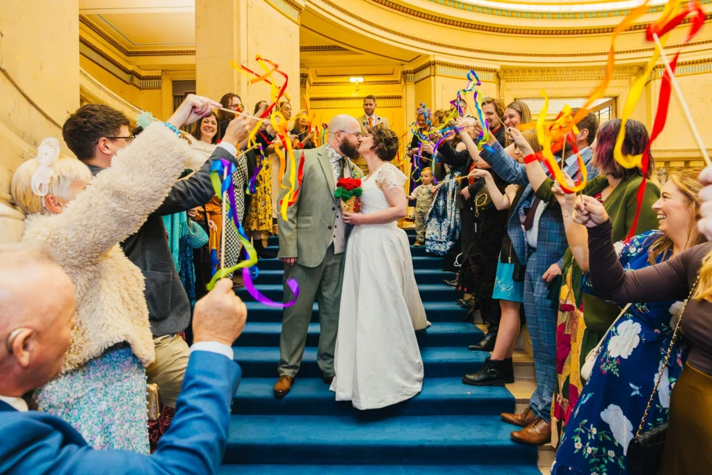 Newlyweds kiss on the blue-carpeted staircase inside Nottingham Council House, surrounded by smiling wedding guests waving colourful ribbon wands.