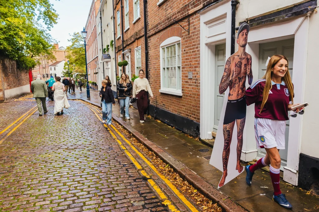 Newlyweds walk down a wet cobbled street in Nottingham as a smiling woman in an Aston Villa kit carries a life-size cardboard cutout of a tattooed man past them.