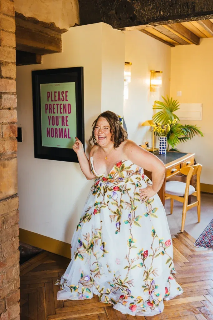 Bride in a colourful embroidered gown laughing and pointing at a Please Pretend You're Normal sign